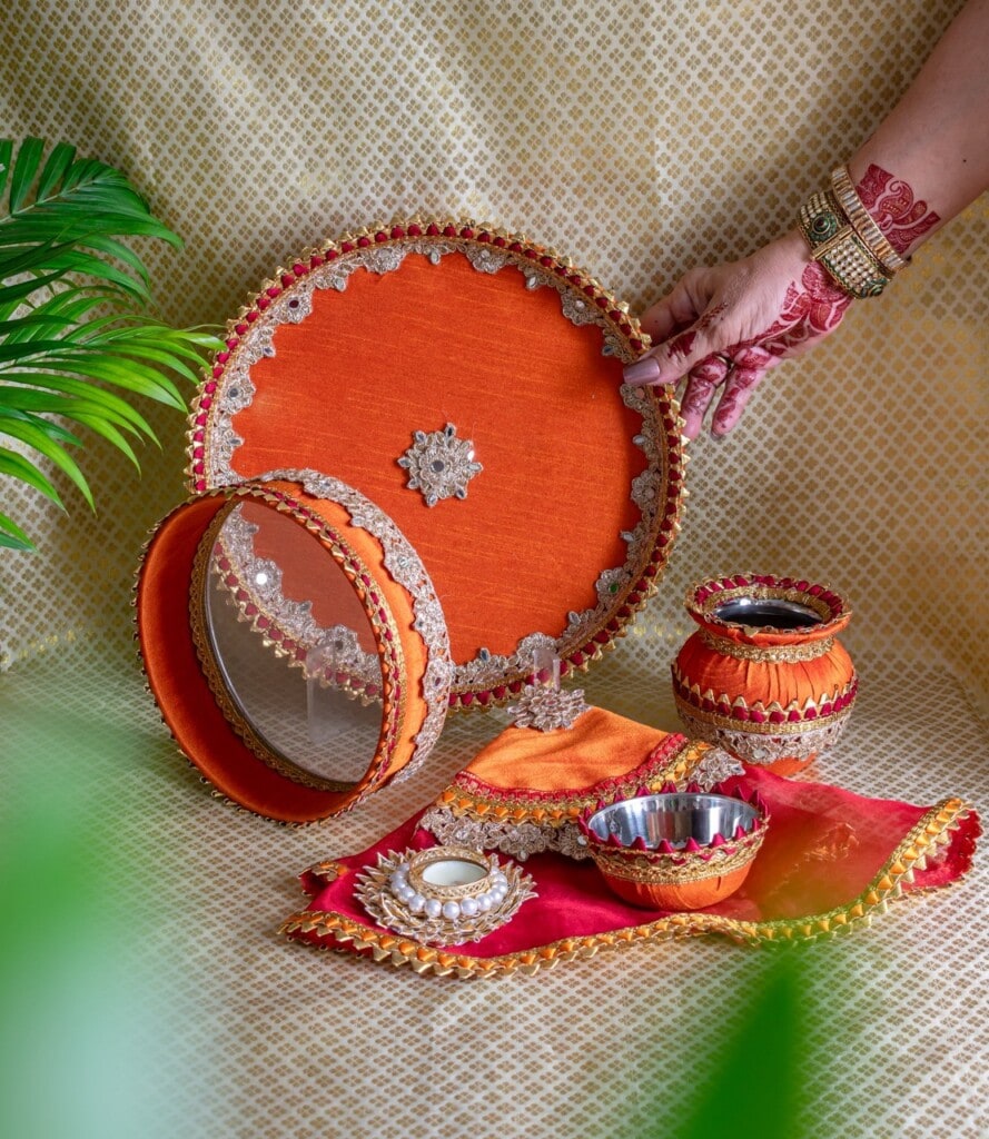 Floral Art presents A decorated Karwa Chauth pooja thali set with a sieve, pot, diya, and cloth, all adorned with orange fabric and gold trim, displayed on a patterned surface.