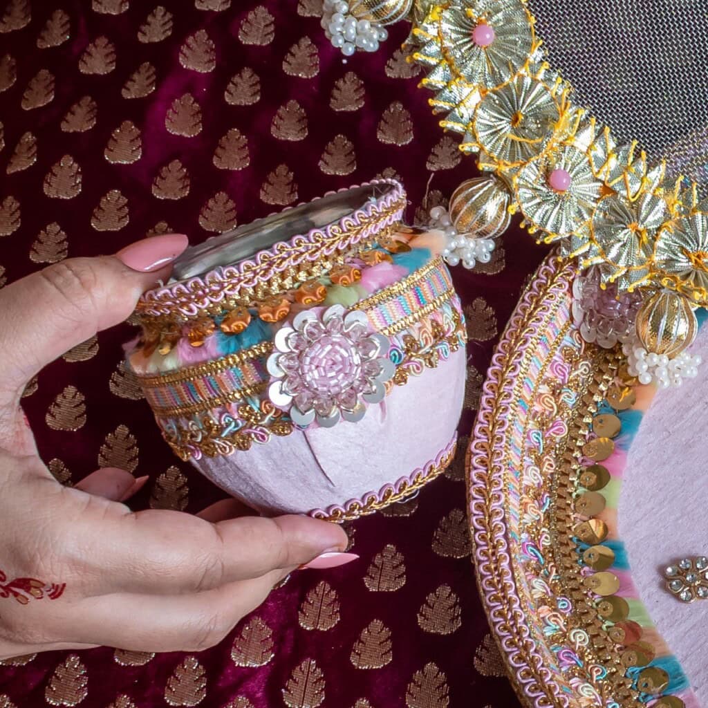 Floral Art presents A hand holding a small, decorative pink pot adorned with intricate gold, silver, and pastel embellishments, alongside a matching tray on a maroon brocade fabric background.