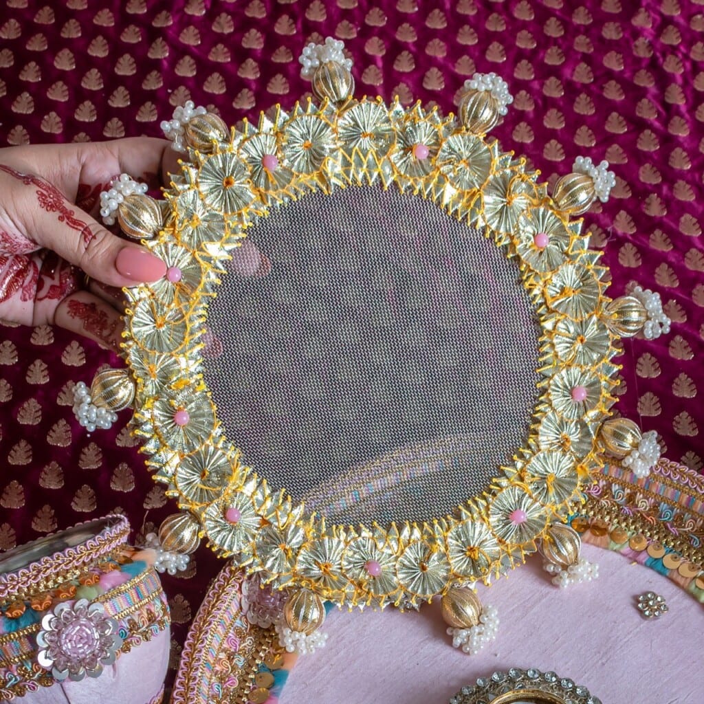 Floral Art presents A hand holds a decorative round sieve with gold and floral embellishments, used for Karva Chauth, against a maroon fabric background with gold motifs.
