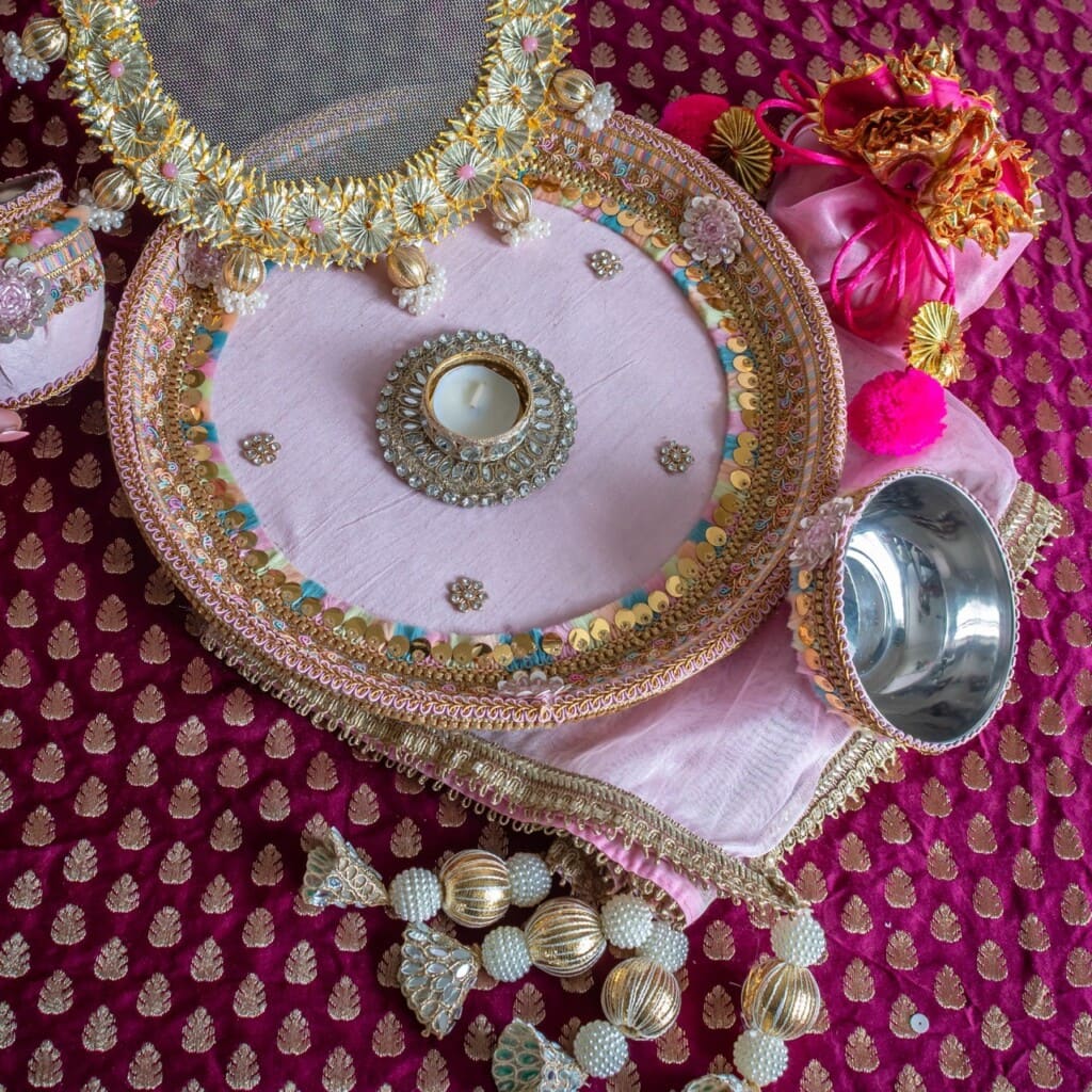 Floral Art presents A decorated thali with a tealight, sieve, cloth, and metal bowl on a pink and gold patterned fabric, accompanied by beaded and pom pom accessories.