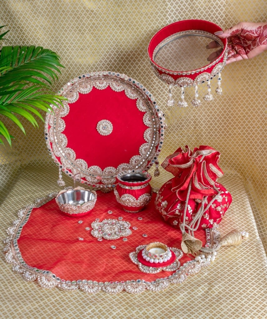 Floral Art presents A decorated red and silver thali set with bowls, a pot, a pouch, and accessories arranged on a patterned surface, with a hand holding a sieve.