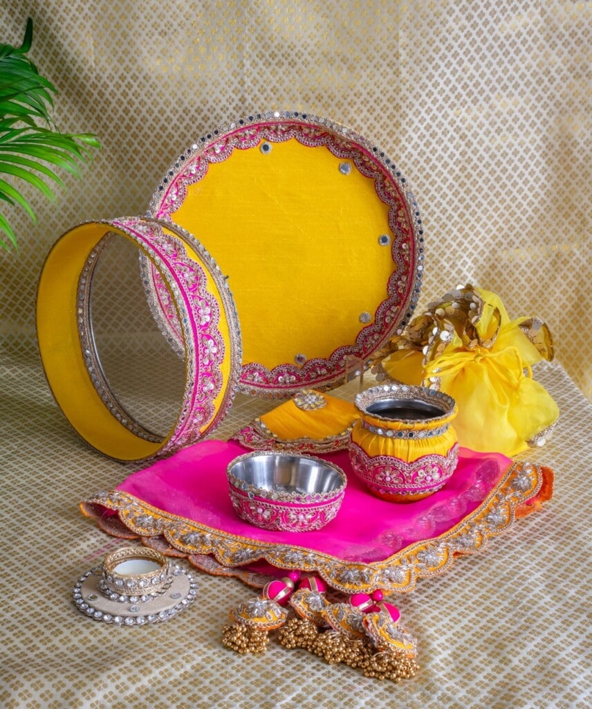Floral Art presents A traditional Indian pooja thali set with decorated trays, bowls, and accessories in yellow and pink, arranged on a patterned fabric background.
