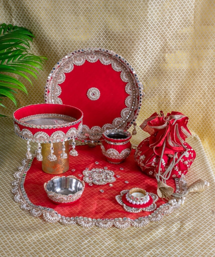 Floral Art presents A decorated red and silver thali set with plates, bowls, and a pouch, arranged on a matching mat with a plant in the background.