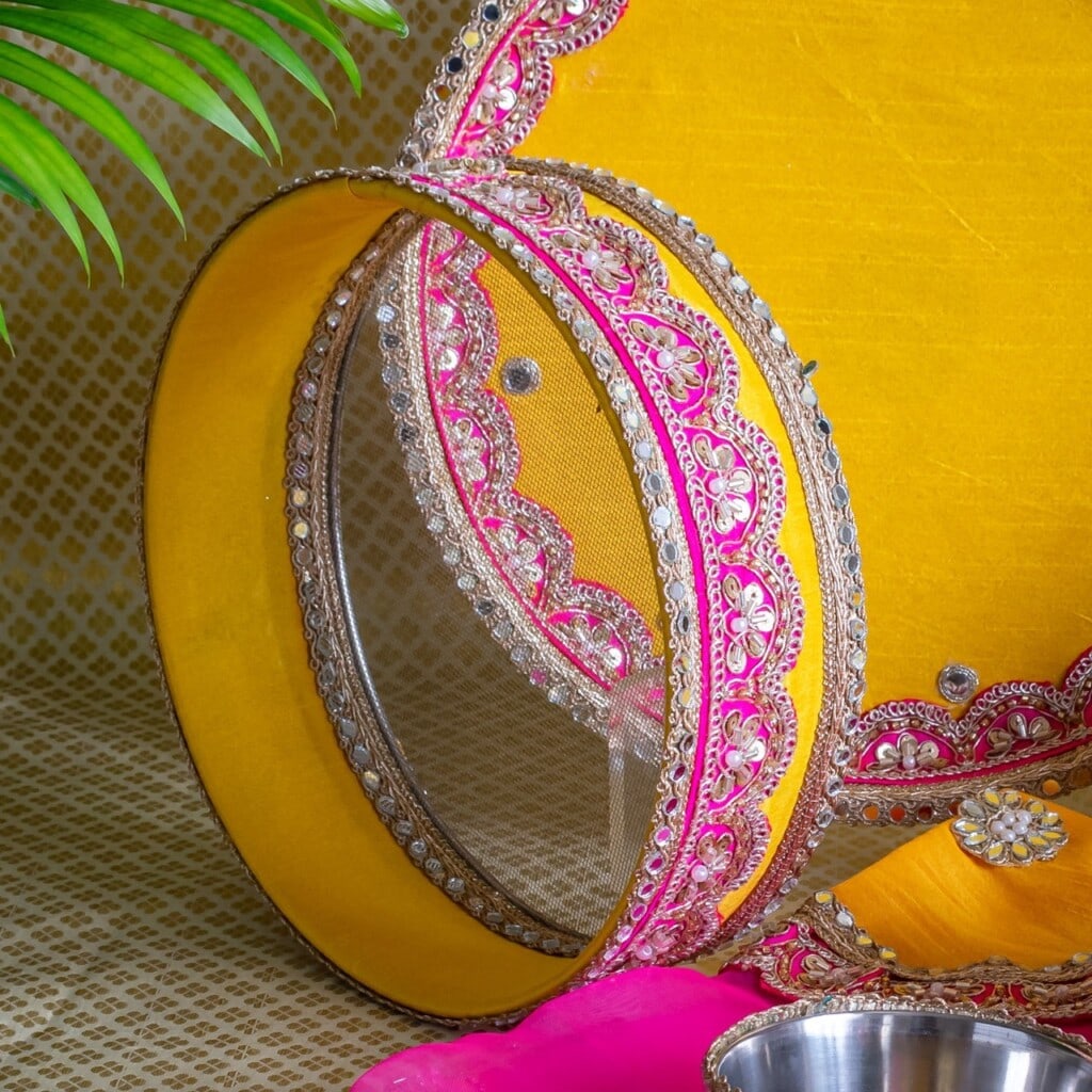 Floral Art presents A decorative yellow and pink sieve with intricate beadwork is displayed upright on a patterned surface with traditional fabric and a leafy plant in the background.