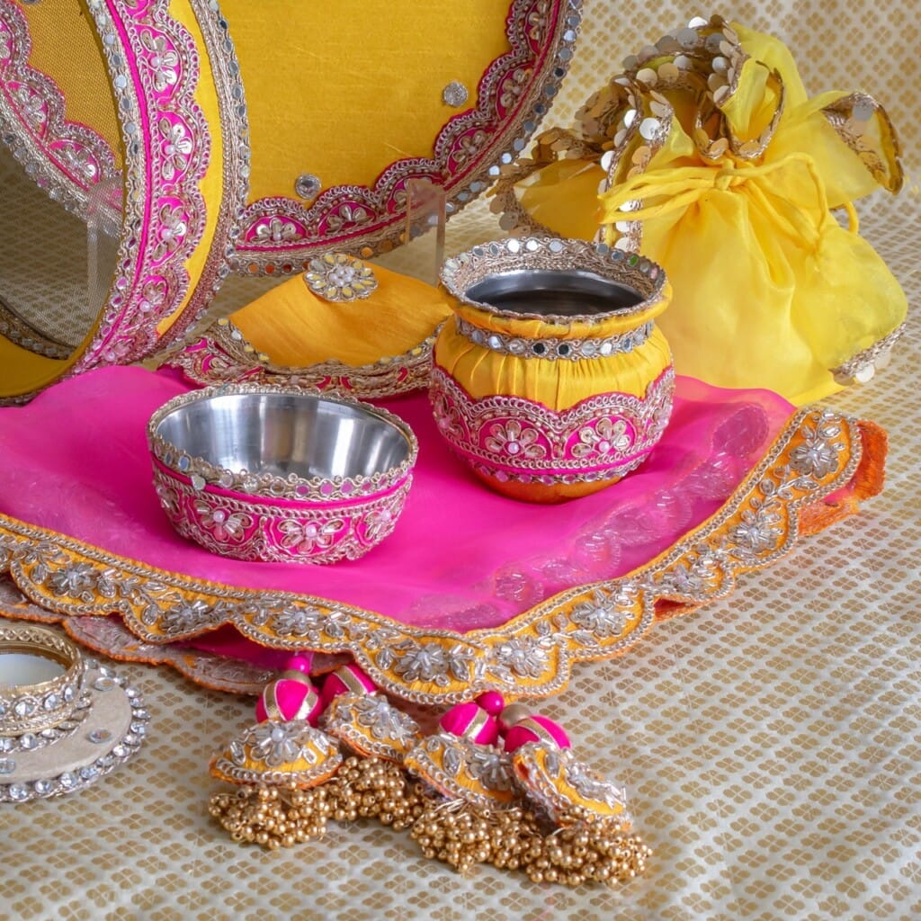 Floral Art presents Traditional Indian puja thali set with decorated tray, bowls, and accessories in yellow and pink fabric on a patterned cloth background.
