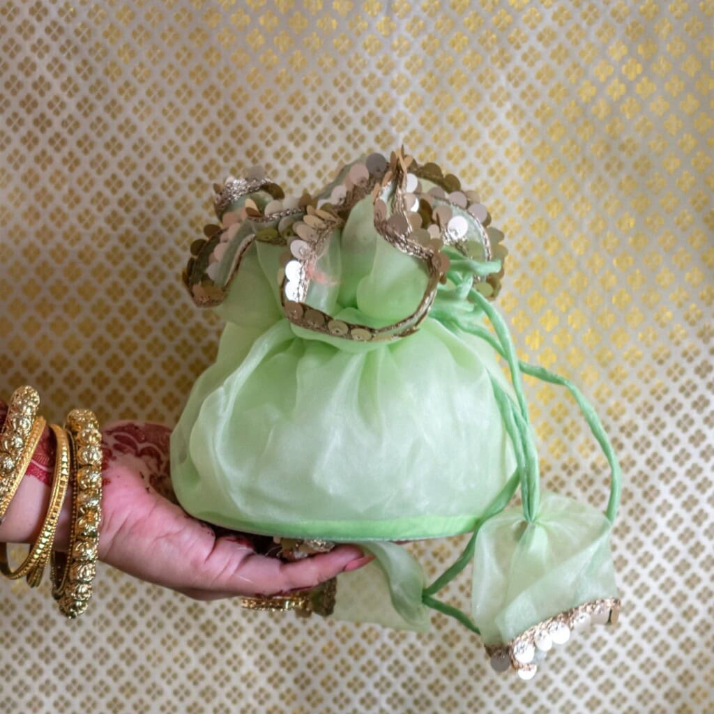 Floral Art presents A hand adorned with gold bangles holds a light green fabric pouch with gold sequin trim, against a patterned background.