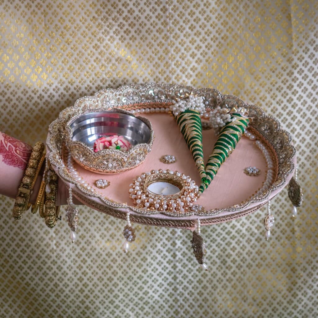 Floral Art presents A decorated thali with a metal bowl, a tealight candle, and two cone shaped green items, held by a person wearing bangles against a patterned golden background.