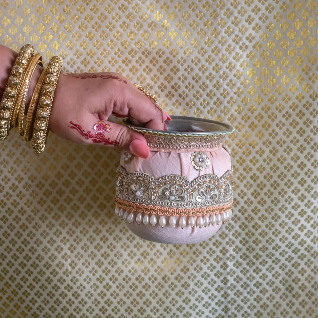 Floral Art presents A hand wearing gold bangles and mehndi holds an ornate decorative pot against a gold patterned background.