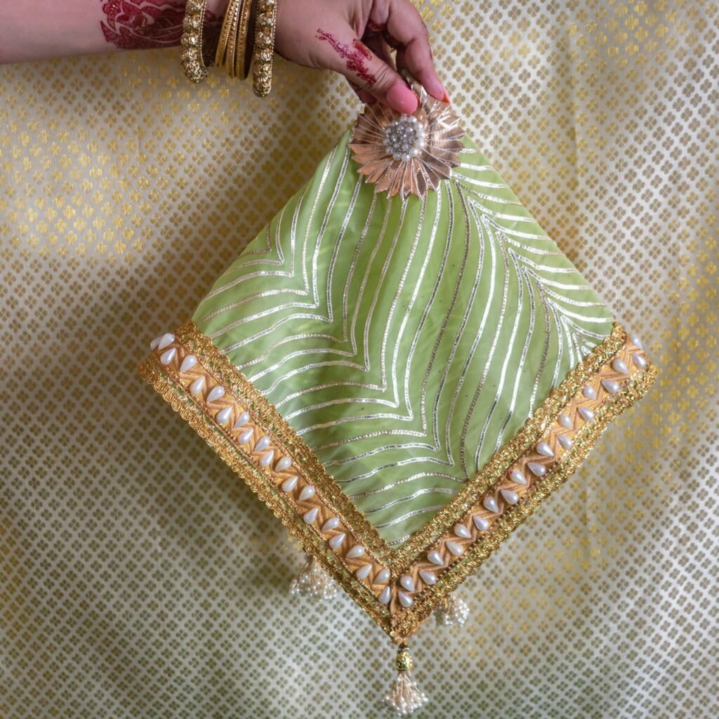 Floral Art presents A hand holds a green decorative potli bag with gold embroidery, shell embellishments, and tassels, against a patterned gold and white background.