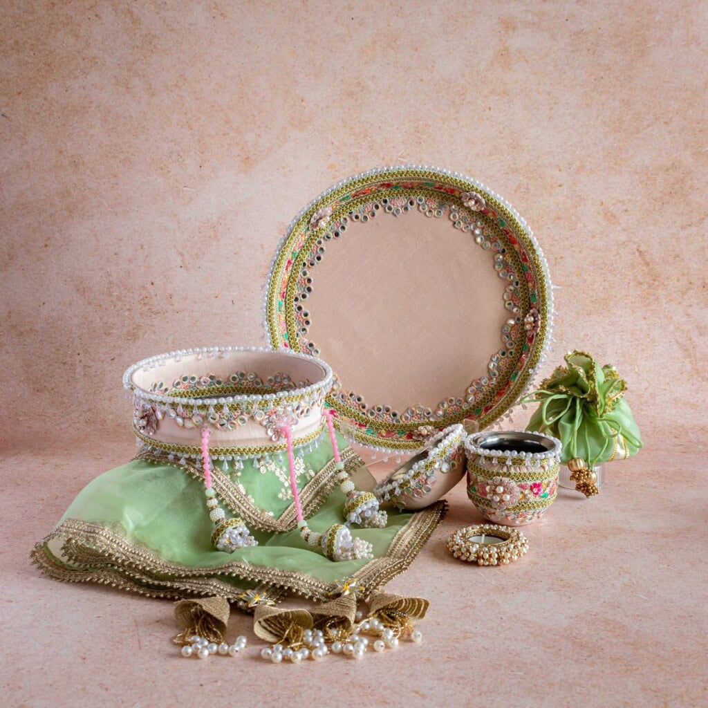 Floral Art presents A set of traditional Indian bridal accessories including a decorated tray, bangles, jewelry, potli bag, and green dupatta with gold trim arranged on a beige background.