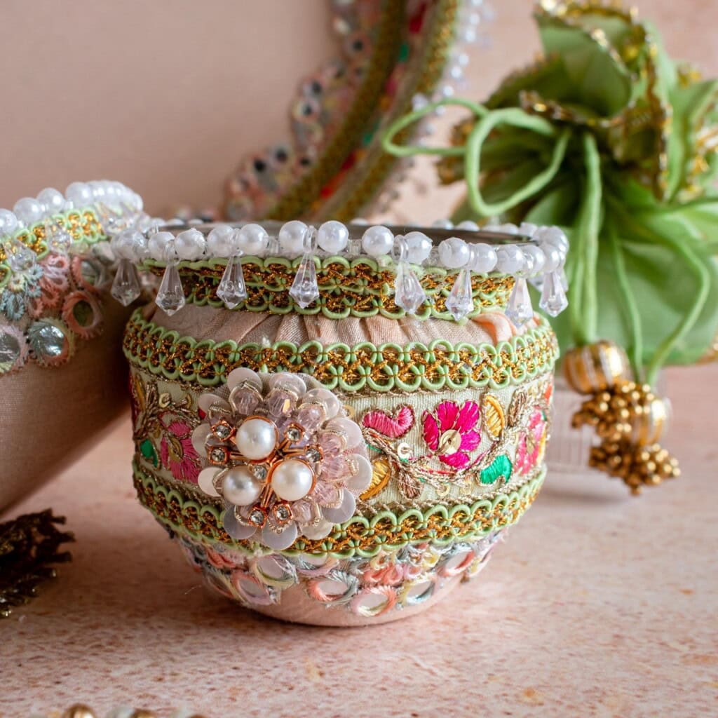 Floral Art presents A decorative pot adorned with pearls, beads, sequins, and floral embroidery, placed on a textured surface with another embellished object in the background.