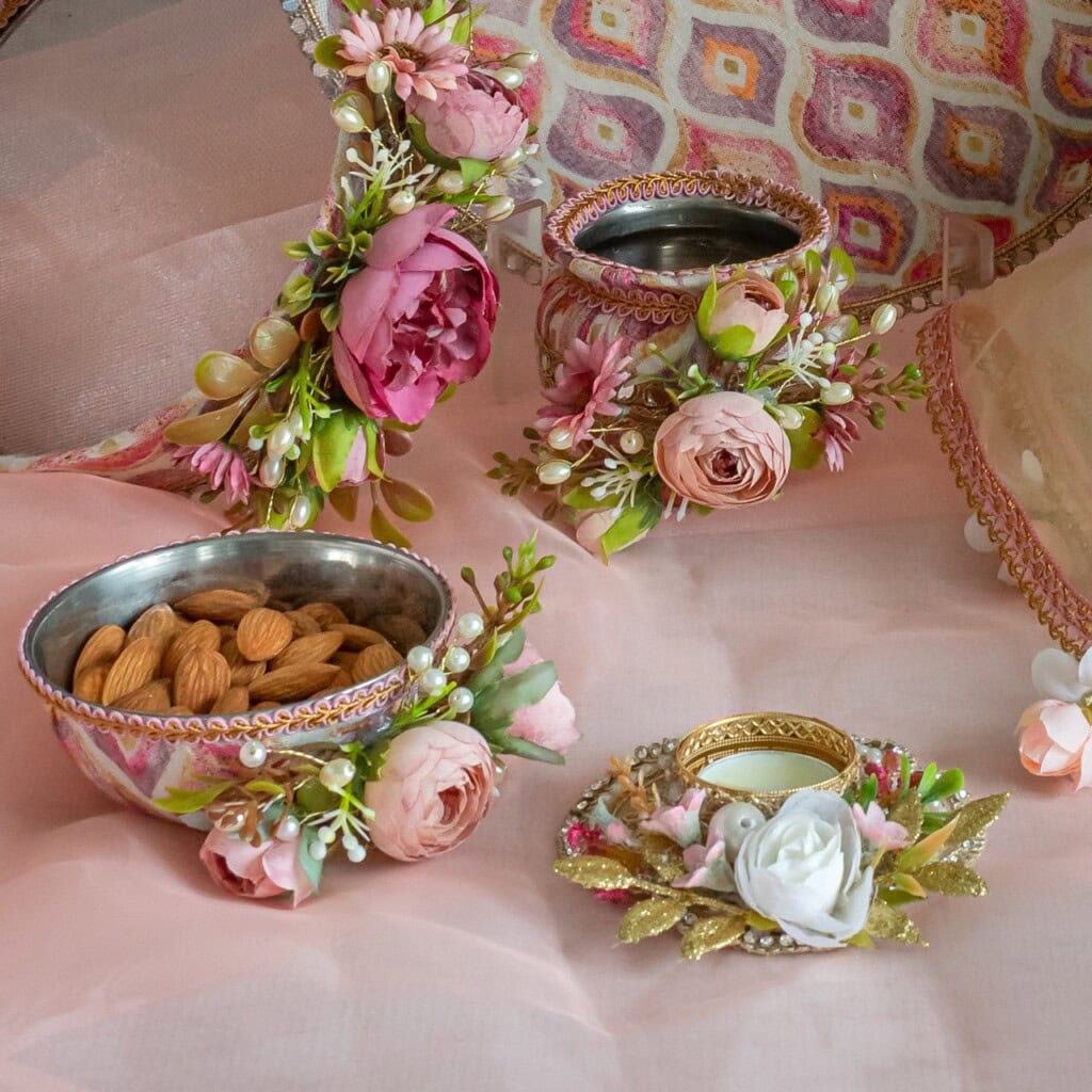 Floral Art presents Decorated bowls with artificial flowers, one filled with almonds, arranged on a pink cloth with a floral patterned textile and a small, embellished candle holder.