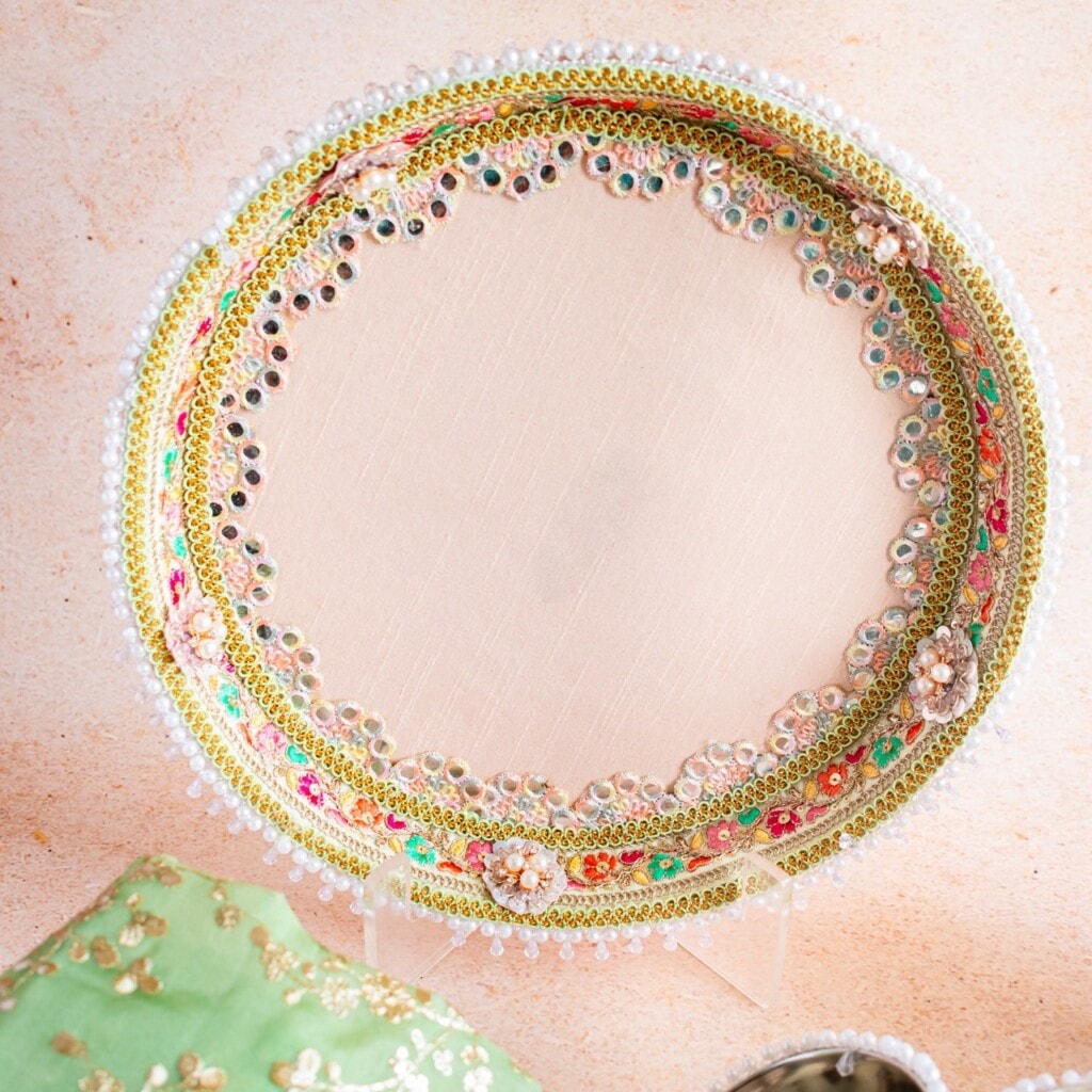 Floral Art presents A round decorative tray with intricate beadwork, embroidery, and mirror details on a light fabric, propped up on a stand against a neutral background.