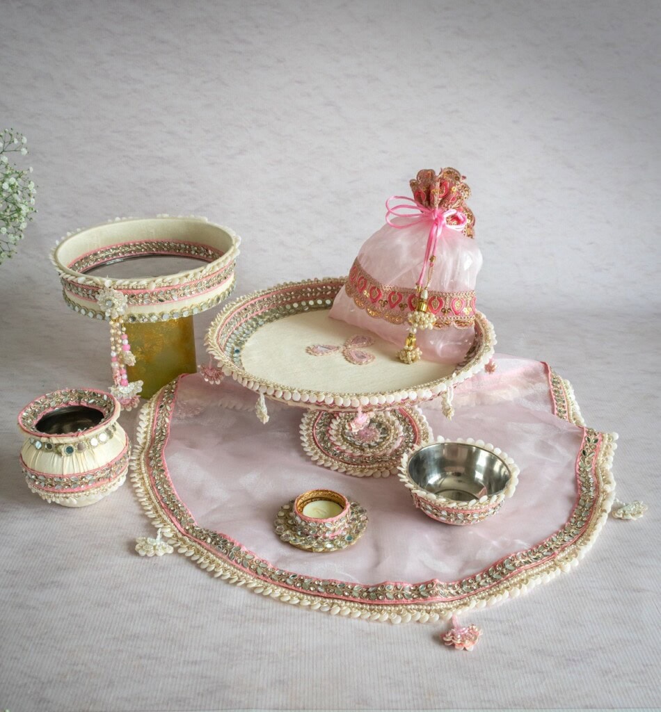 Floral Art presents Traditional Indian pooja thali set with decorated plates, bowls, a diya, and a pot, all adorned with pink and gold embellishments, arranged on a light background.