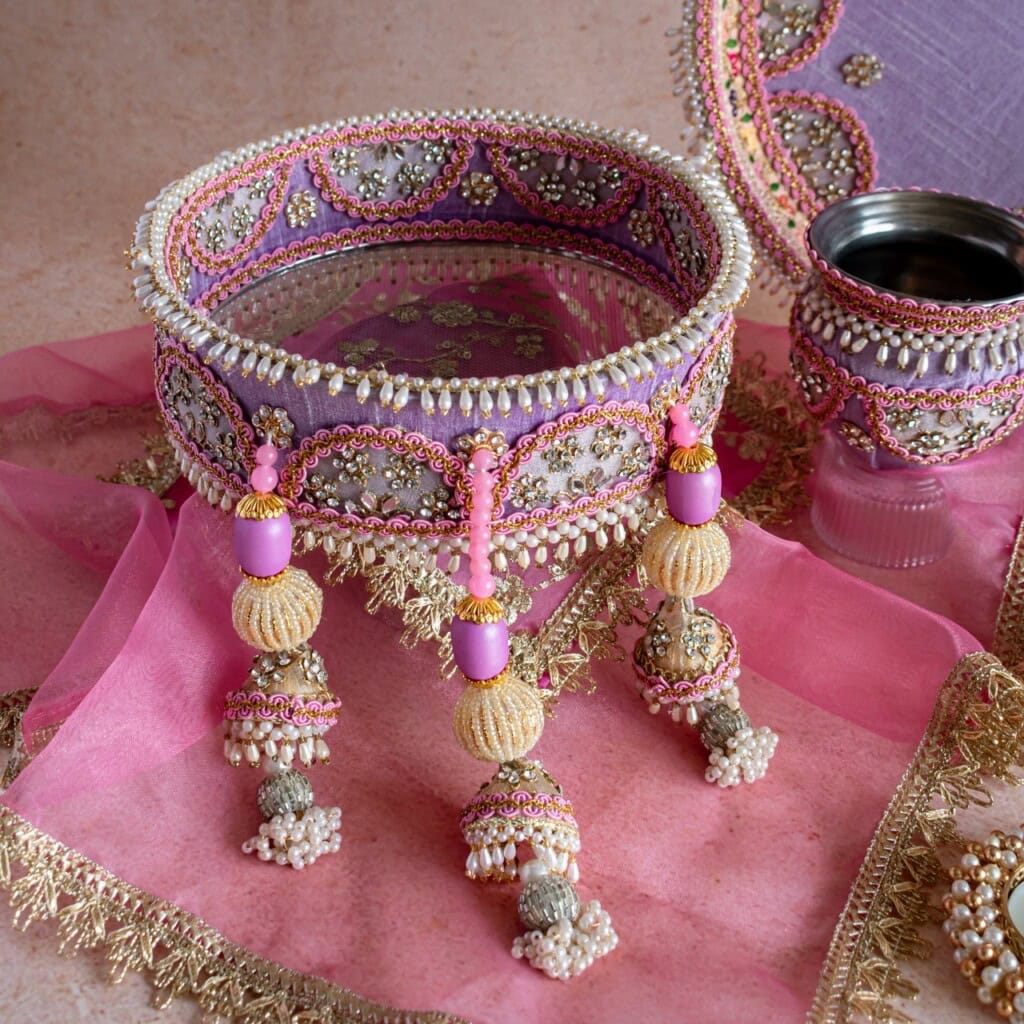 Floral Art presents A decorated sieve with beads and embroidery, a glass, and a pink cloth with gold trim, arranged on a light surface.