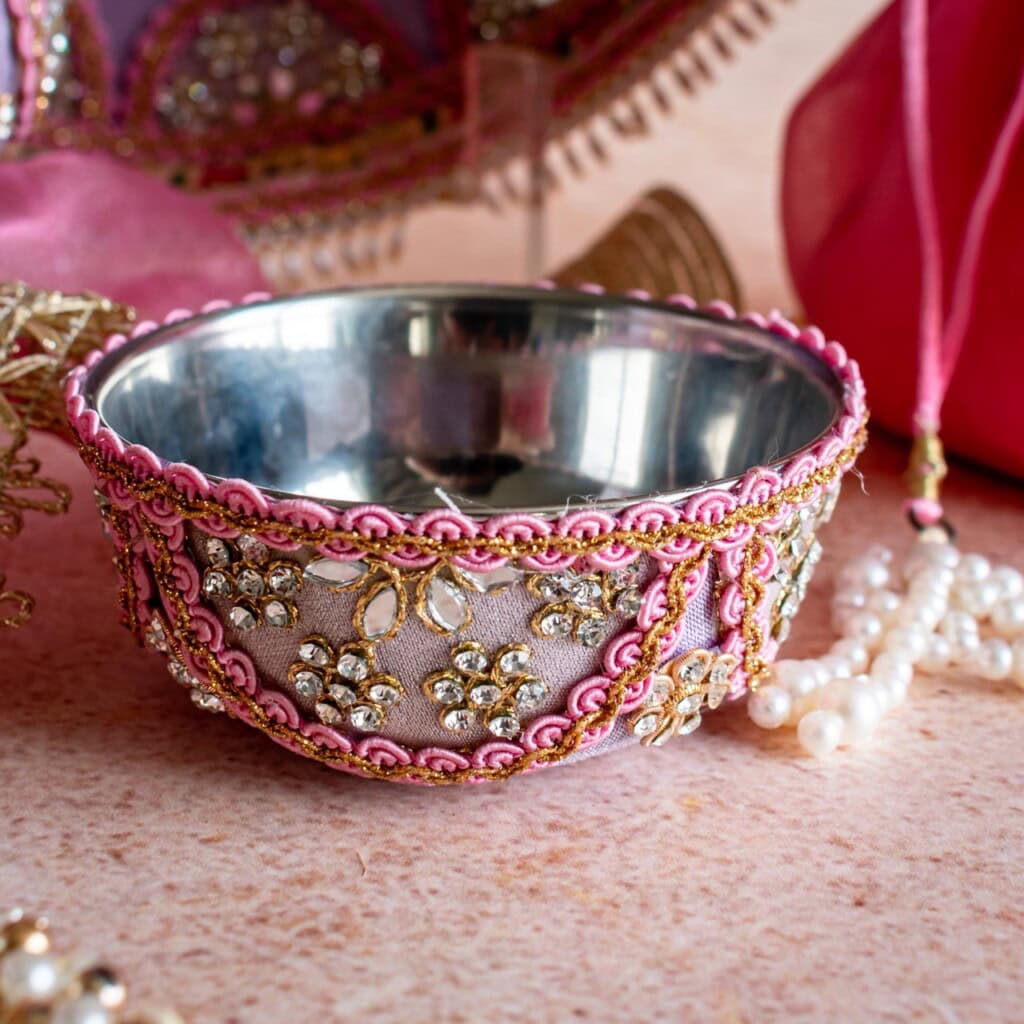 Floral Art presents A stainless steel bowl decorated with pink fabric, gold embroidery, and white beads, placed on a light pink surface with pearl jewelry nearby.