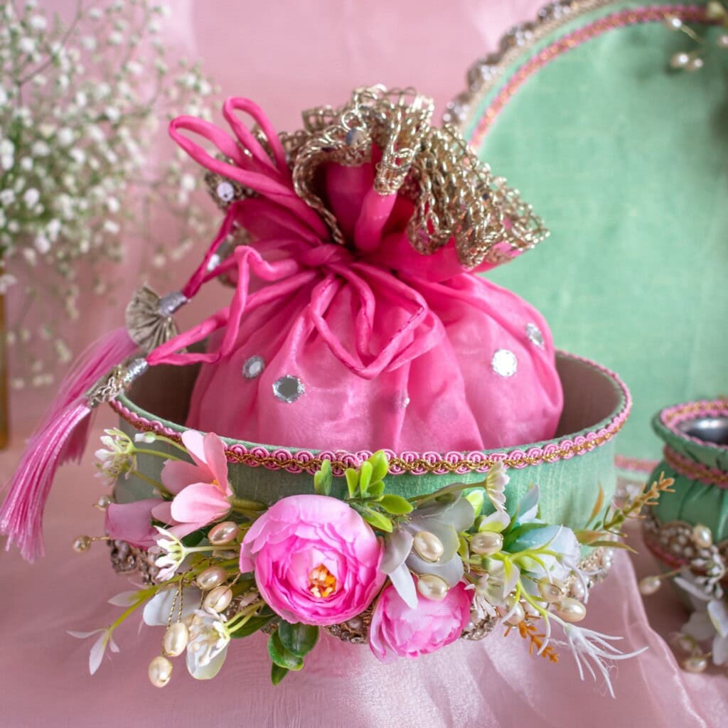 Floral Art presents A pink drawstring pouch with decorative silver trim sits in a green and pink basket adorned with artificial flowers and pearls.