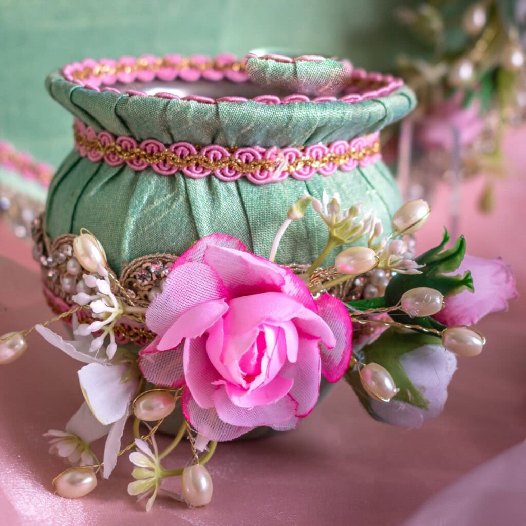 Floral Art presents A decorative green fabric jar with gold and pink trim, adorned with artificial pink roses, white buds, and beads, set against a soft, pastel background.