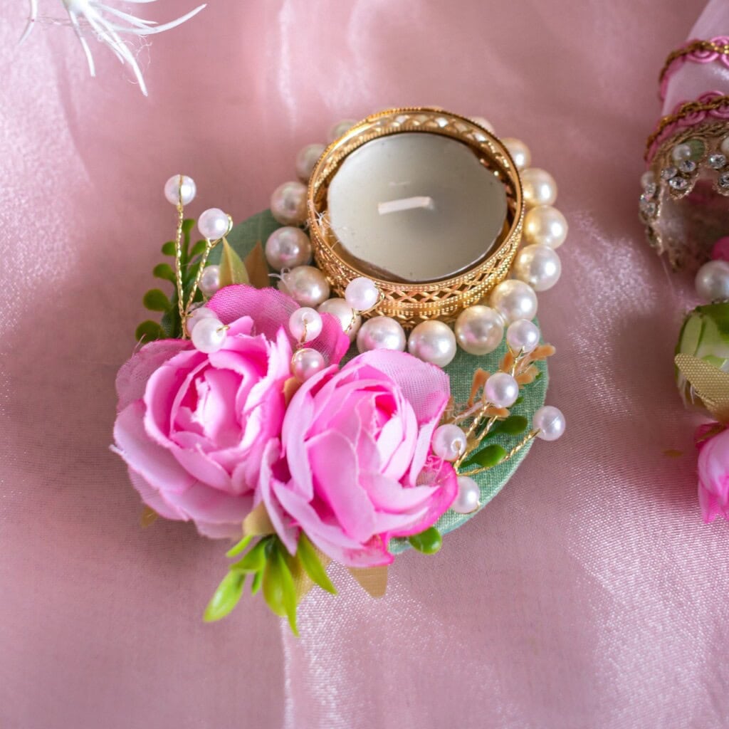 Floral Art presents A decorative tealight candle holder adorned with faux pink roses, green leaves, white pearls, and a gold rim sits on a pink satin fabric.