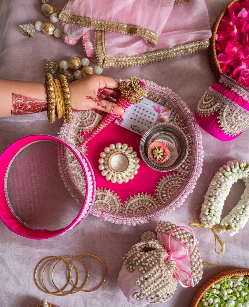 Floral Art presents A woman with henna on her hand holds a decorated sieve over a pink and gold thali containing a candle, water, and a red container. Traditional jewelry and festive items surround the thali.