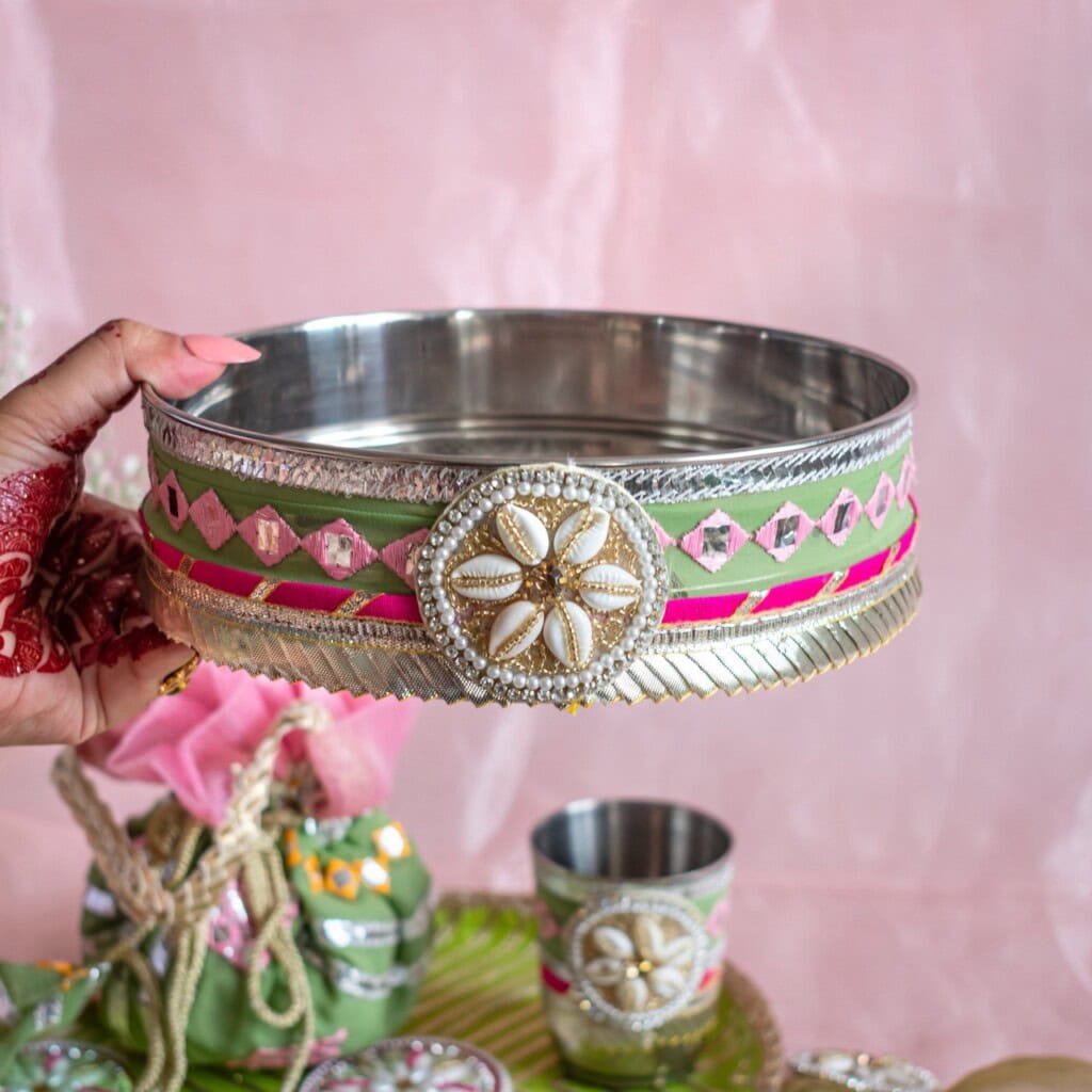 Floral Art presents A hand holds a decorated metal bowl adorned with beads, shells, and pink and gold ribbons, with matching items in the background.