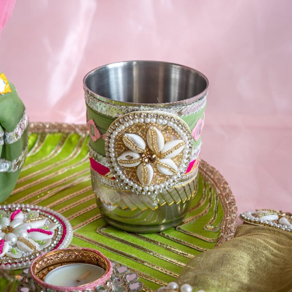 Floral Art presents A stainless steel cup decorated with beads, shells, and ribbons sits on a green and gold tray, surrounded by other festive items.