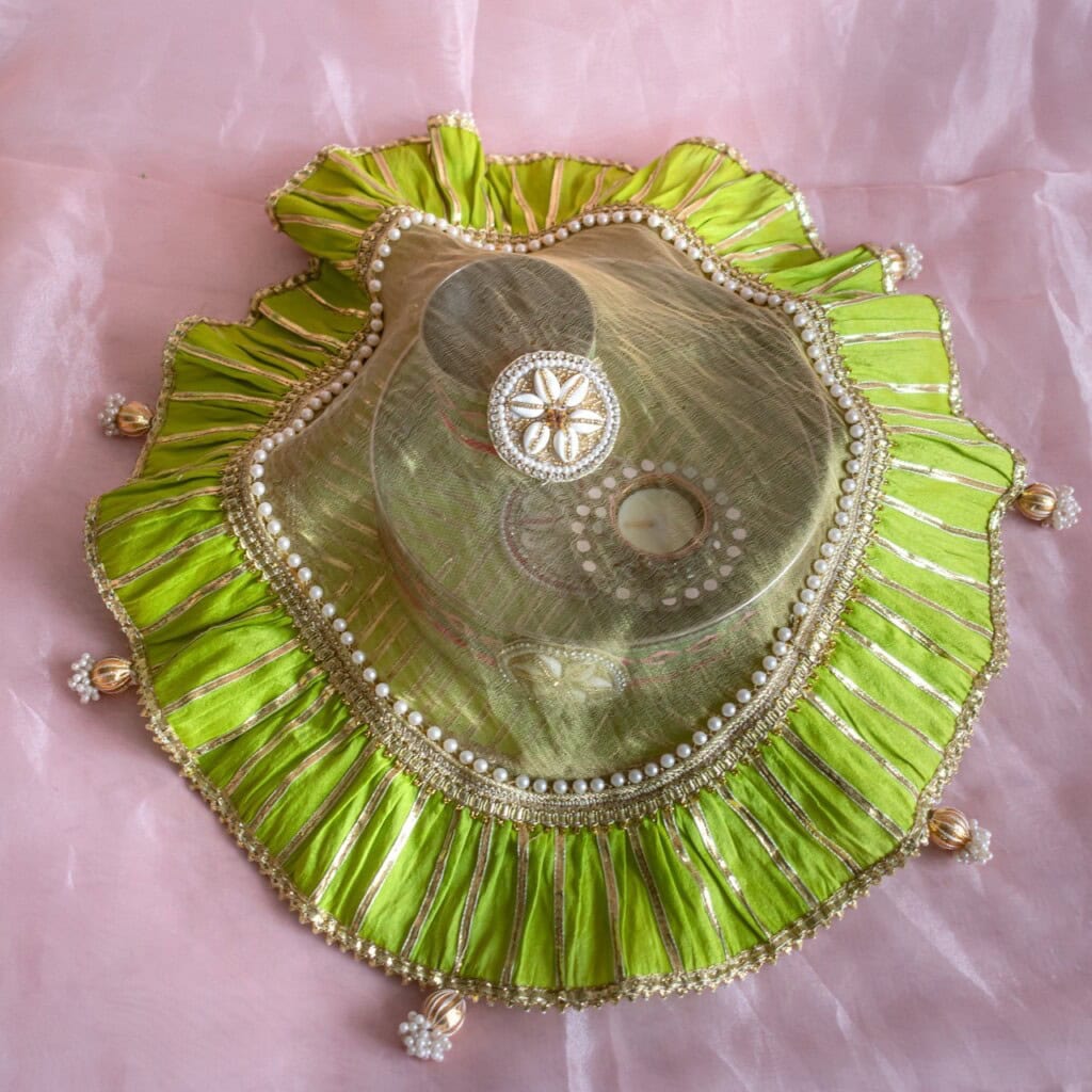 Floral Art presents A decorative green and gold fabric cover with beads and pleated edges, placed on a pink background.