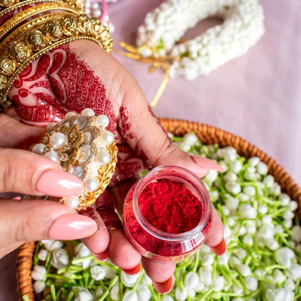 Floral Art presents A hand with traditional jewelry and henna holds a small jar of red sindoor powder above a basket of white flowers and a floral garland.