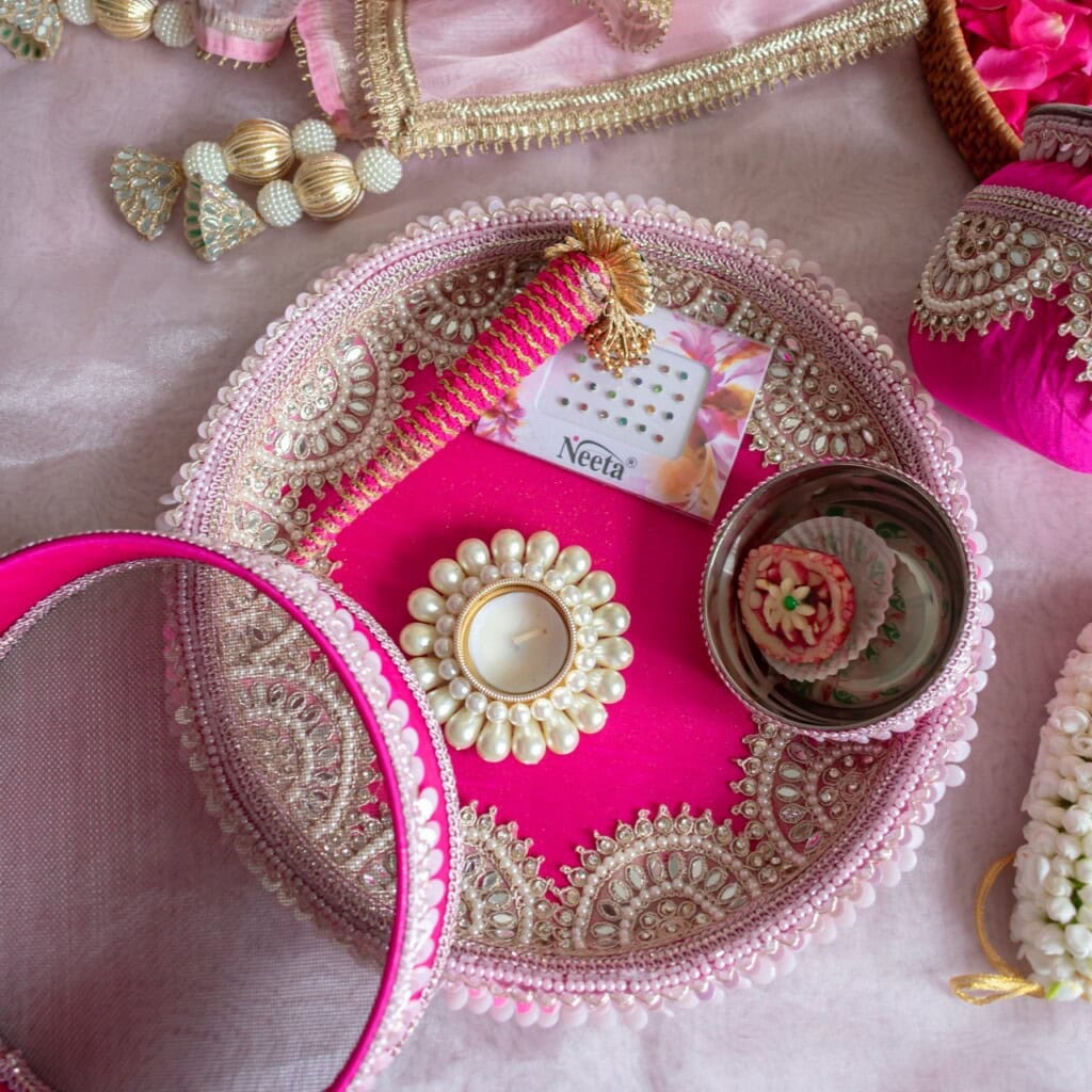 Floral Art presents A decorated pink thali with a pearl candle holder, traditional jewelry, a comb, a small bowl with sweets, and a pink cloth, used for an Indian celebration.