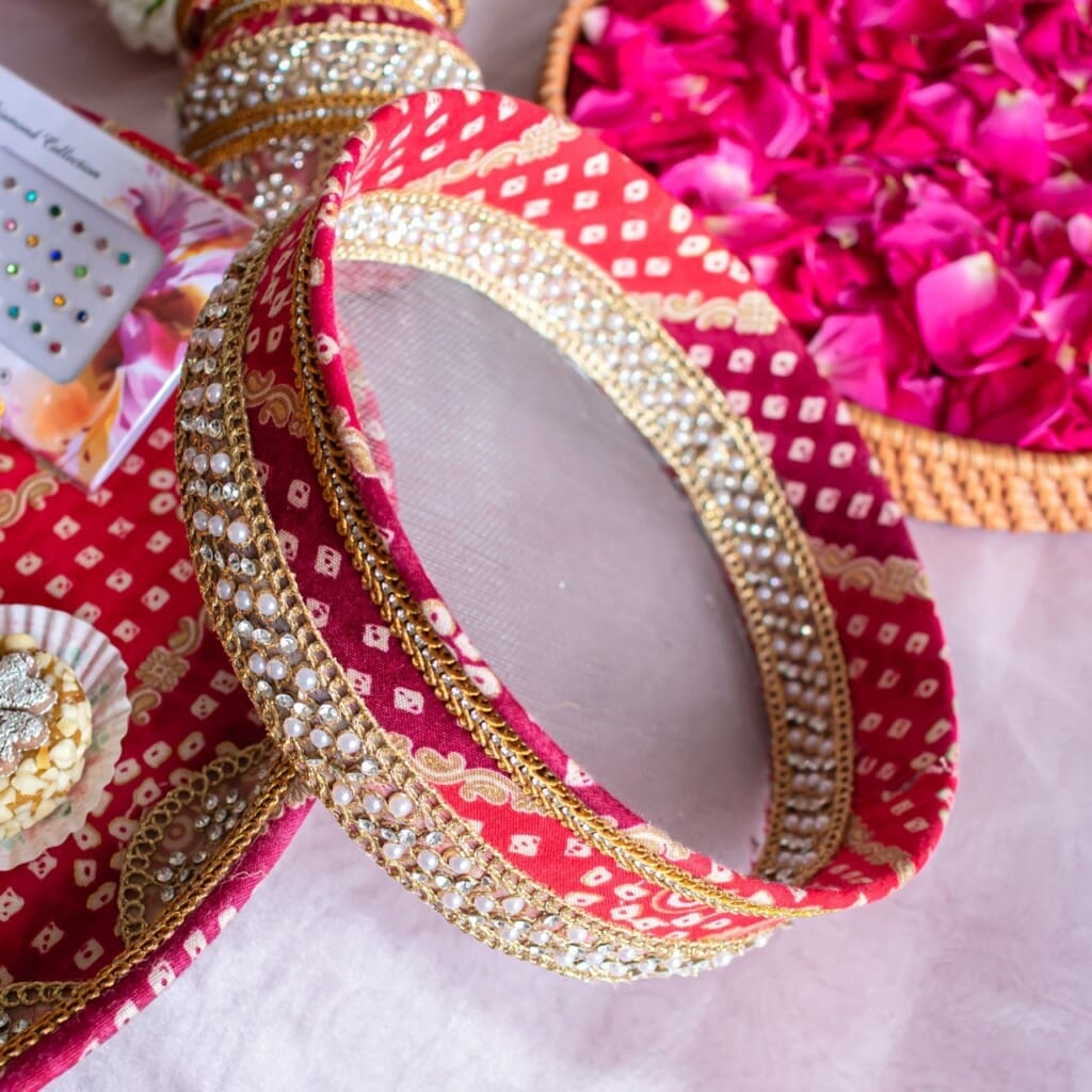 Floral Art presents A decorated red and gold sieve with embroidery and beads, used in Indian festivals, placed beside rose petals and traditional accessories on a table.