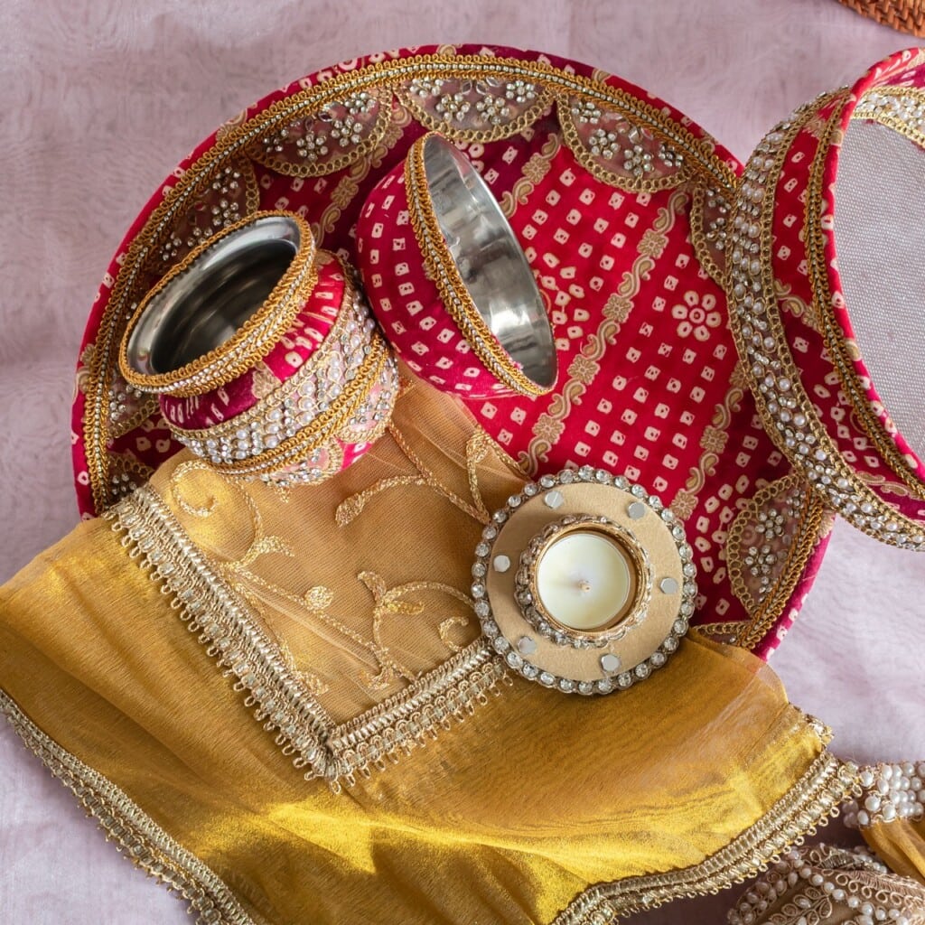 Floral Art presents A decorated Karva Chauth thali set with a steel tumbler, sieve, and candle placed on a red and gold embroidered cloth.