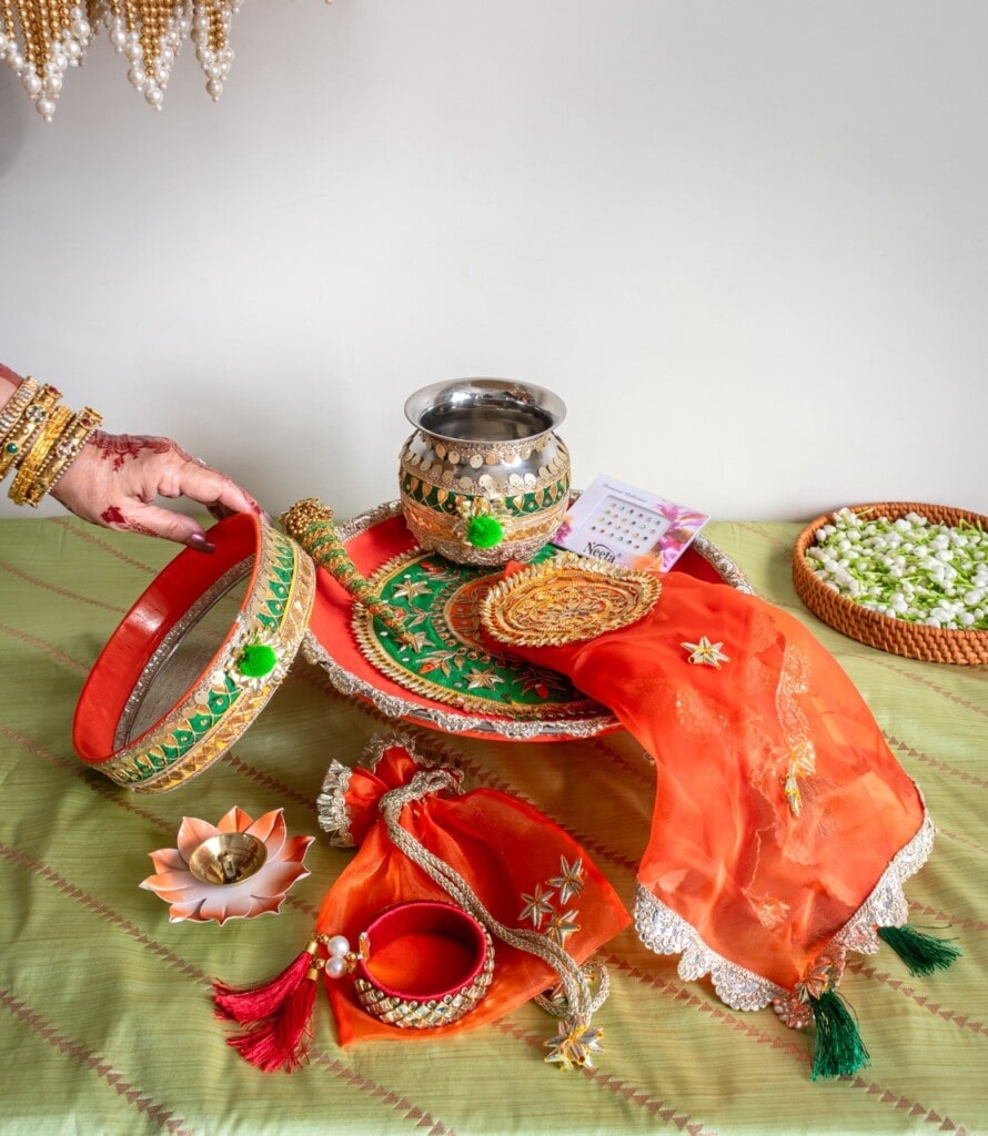 Floral Art presents A decorated thali with traditional items for Karva Chauth, including a sieve, kalash, bangles, a diya, and red fabric on a green cloth.
