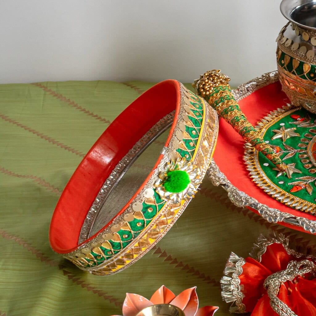 Floral Art presents A decorated sieve with green and gold embellishments placed on a green cloth, next to a traditional vessel and festive items.