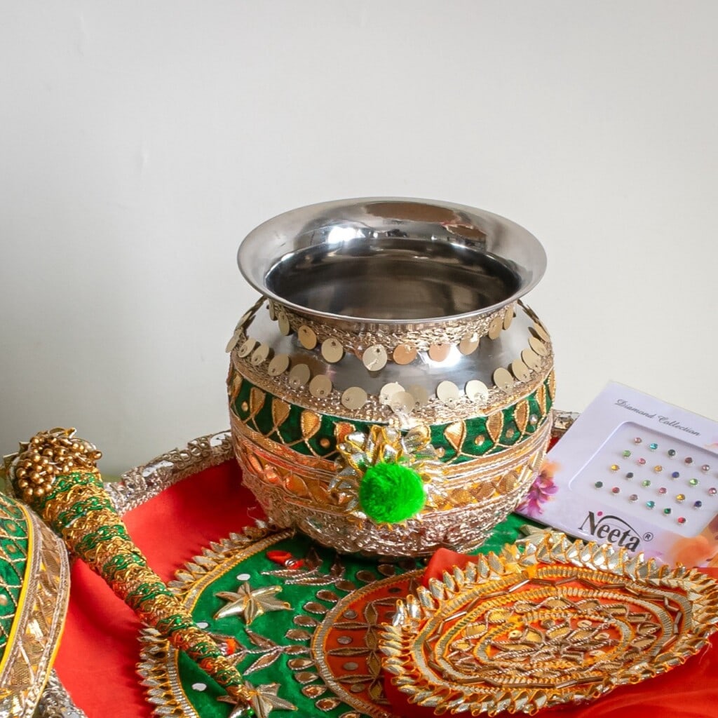 Floral Art presents A decorated silver pot is placed on a red and green cloth with gold embroidery, accompanied by decorative accessories and a packet of bindis.