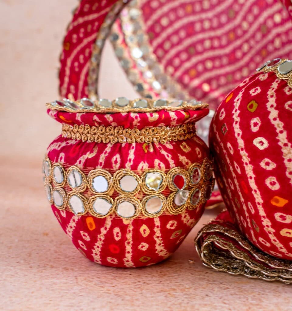 Floral Art presents A decorative red pot adorned with gold trim and mirror work, placed against a patterned red and gold fabric background.