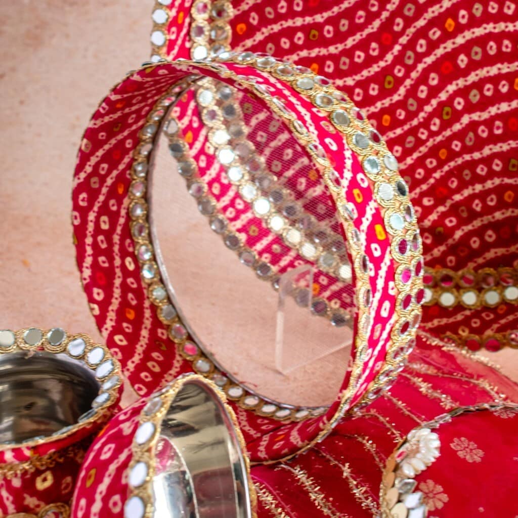 Floral Art presents A decorated red and gold sieve with mirrors, used for Karwa Chauth, is placed alongside matching bowls and a red embroidered cloth.