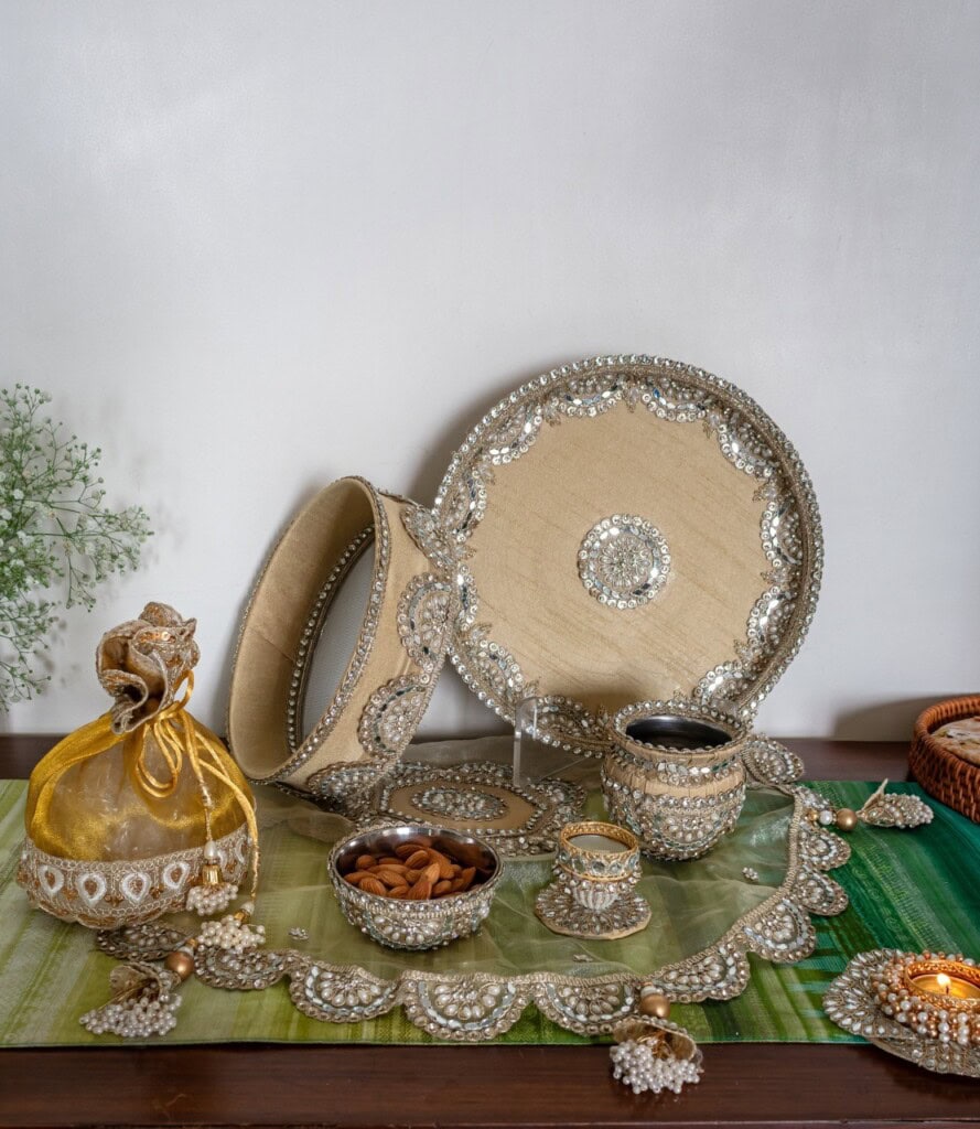 Floral Art presents A decorative Karwa Chauth thali set with beaded embellishments, including a tray, sieve, cups, sweets, and a pouch, arranged on a green cloth.