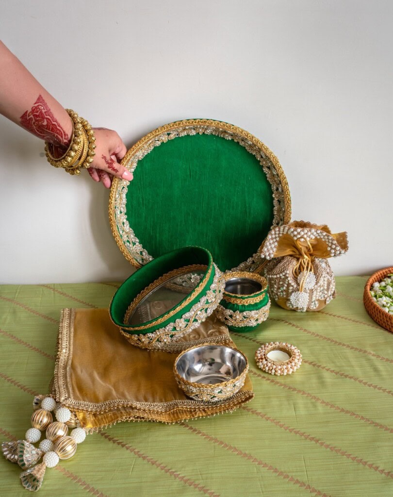 Floral Art presents A hand wearing gold bangles and henna holds a decorated green tray, with matching bowls, a pouch, and a cloth arranged on a green surface.