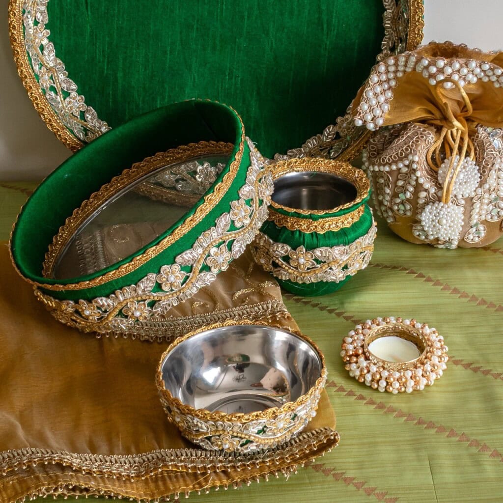 Floral Art presents Traditional decorative trays, bowls, a pouch, and a tealight holder with gold, green, and pearl embellishments are arranged on a green and brown fabric background.