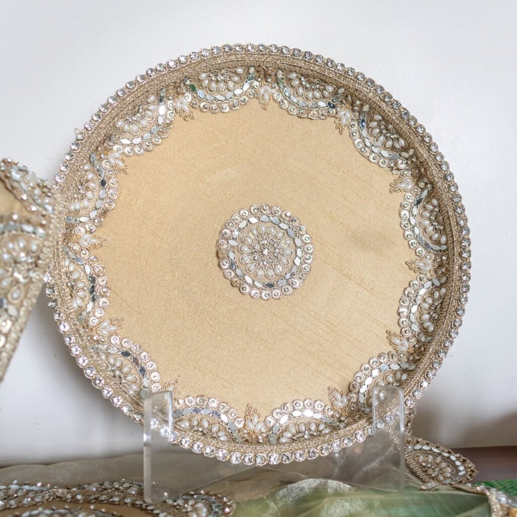 Floral Art presents A round, gold decorative tray with intricate bead and sequin designs along the edge and center, displayed upright on a clear stand.