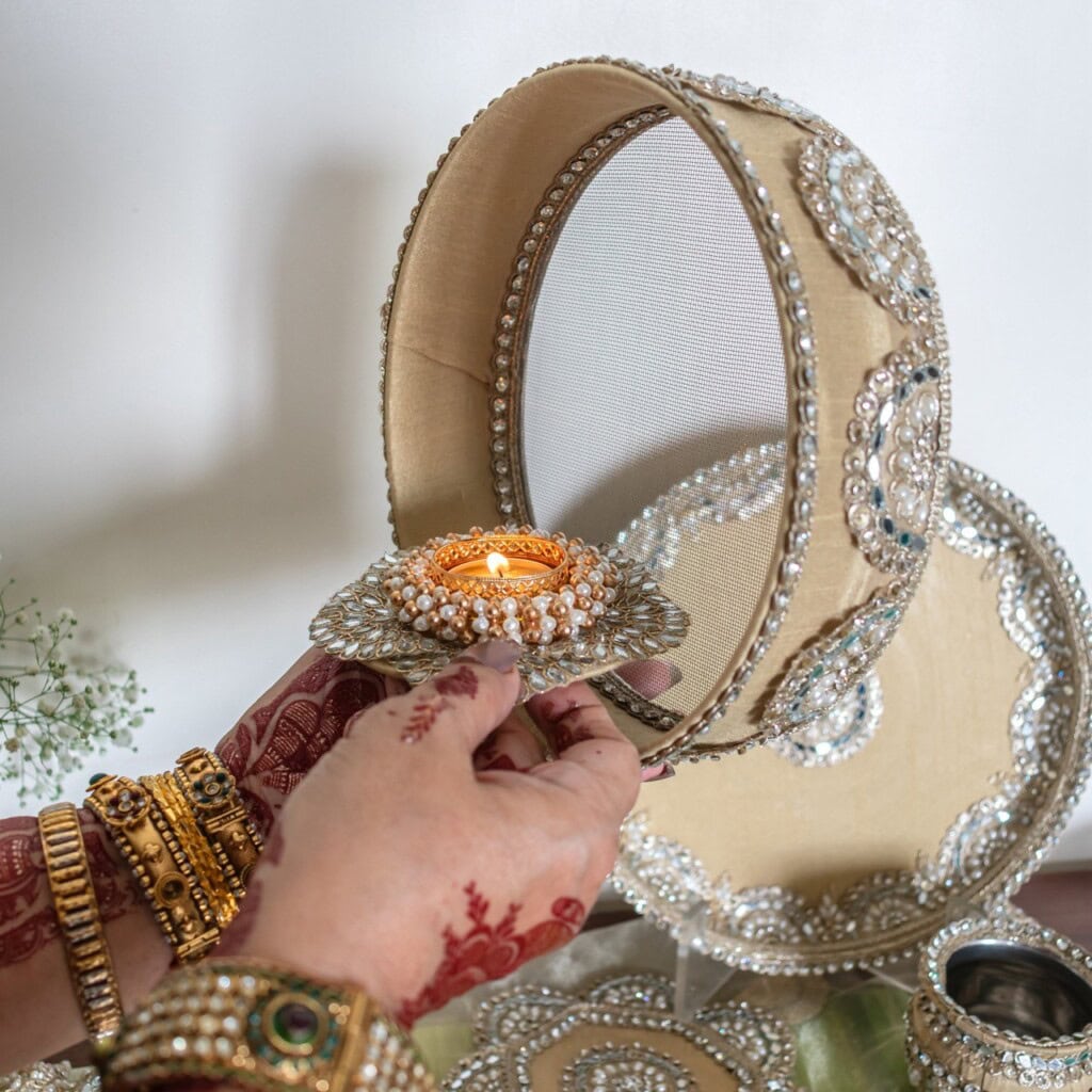 Floral Art presents A decorated sieve and plate set is held by a woman with henna and jewelry, as she lights a candle, preparing for a traditional ritual.