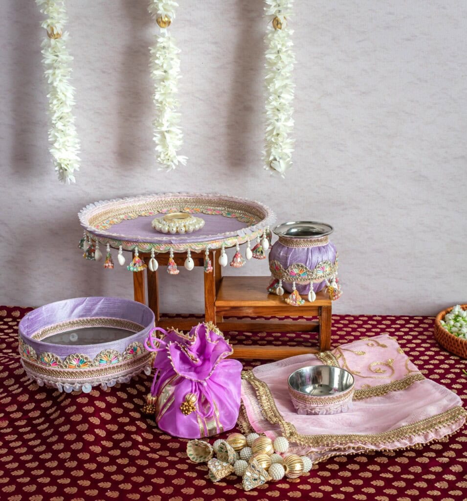 Floral Art presents Decorative Indian pooja thali set with bowls, cloth, and pouch arranged on a maroon patterned surface with white flower garlands hanging in the background.