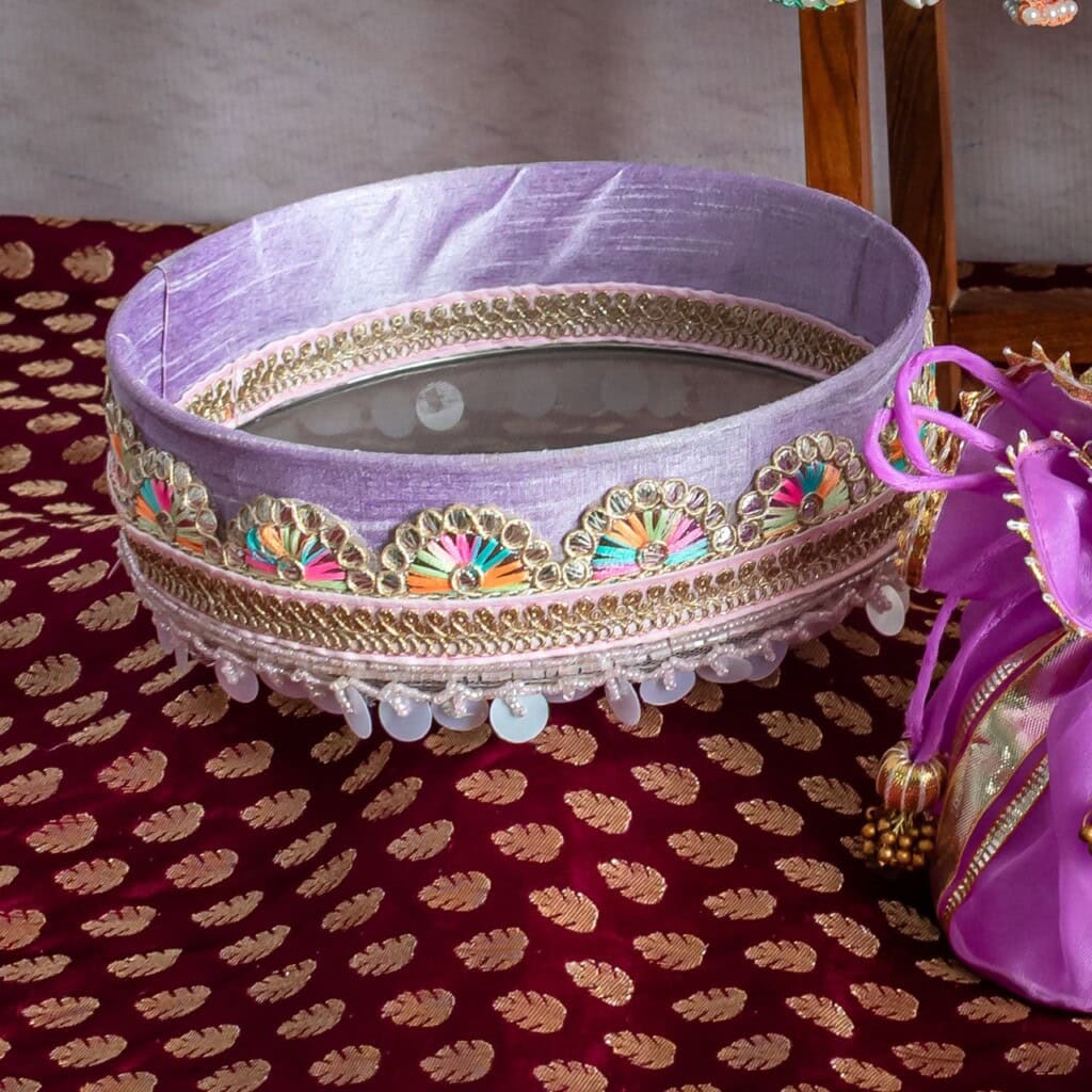 Floral Art presents A decorated, round sieve with a mirror at the base, adorned with purple fabric, beads, and embroidery, placed on a maroon patterned cloth.