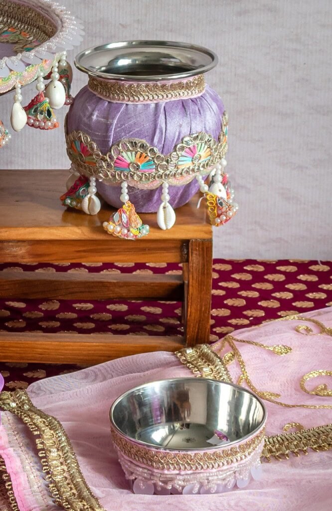 Floral Art presents A decorative metal pot covered in purple cloth with beads and embellishments sits on a wooden table next to a matching metal bowl and pink embroidered fabric.